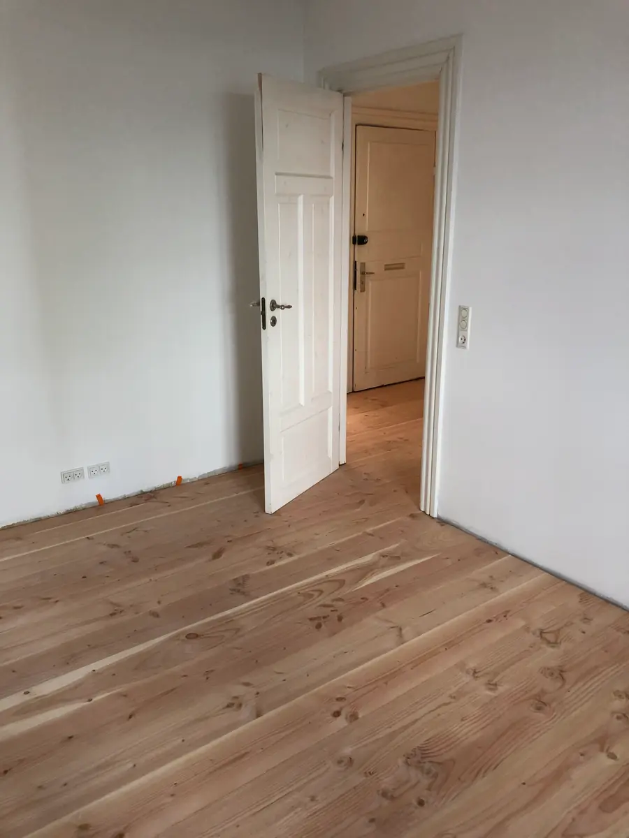 Ubehandlet fyrgulv gennem døråbning med hvid fyldingsdør åben mod entré, hvide vægge og afstandskiler ved fodpanelet
