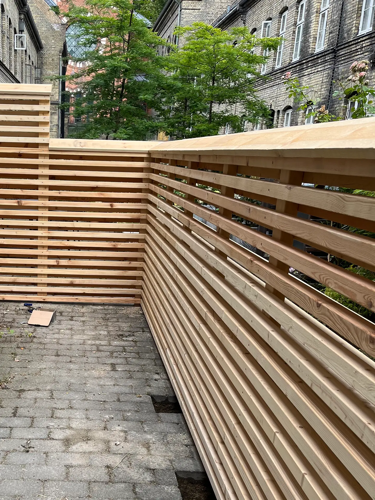 Lamelhegn i lyst, ubehandlet træ danner hjørne i københavnsk gård med gulstensfacader og gråt fliseareal