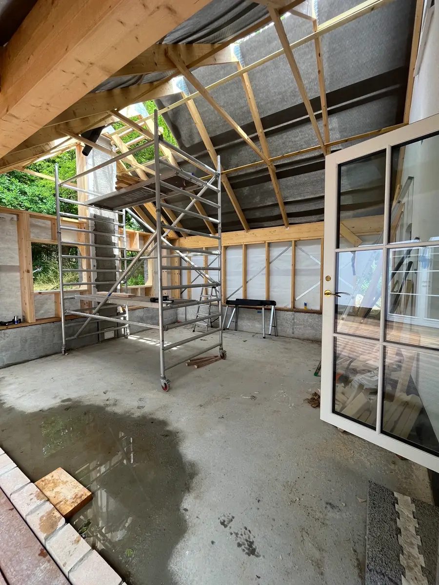 Råhuset indefra med stolpevægge, undertag på spærene, rullestillads på gulvet og hvid sprossedør mod haven