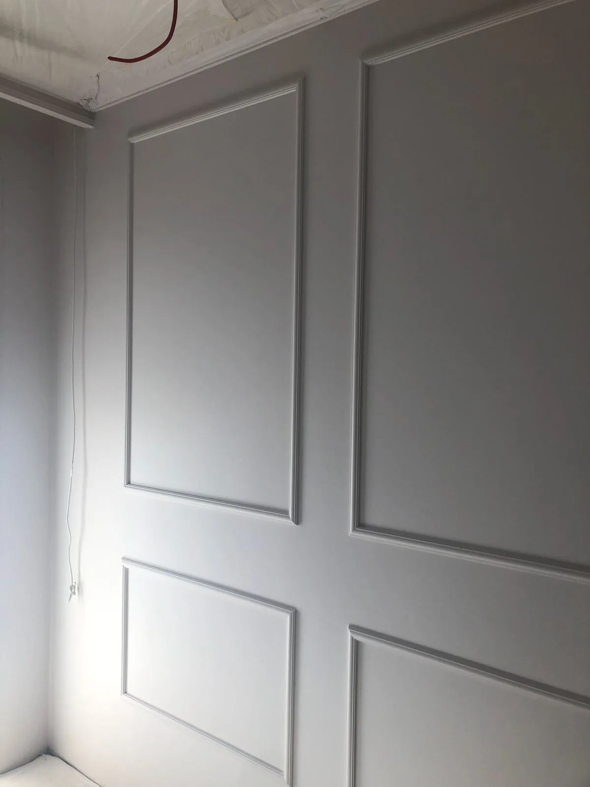 Væg med klassiske panelrammer i to rækker, spartlet og malet i én lysegrå tone, med en løs kabelende hængende fra det ubehandlede loft i hjørnet